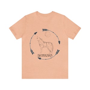 Puede incluir: Una camiseta de color melocot&oacute;n con una ilustraci&oacute;n en blanco y negro de un lobo aullando a la luna. El lobo est&aacute; rodeado por un c&iacute;rculo de flechas con la palabra "SHUNKARA" escrita dentro del c&iacute;rculo.