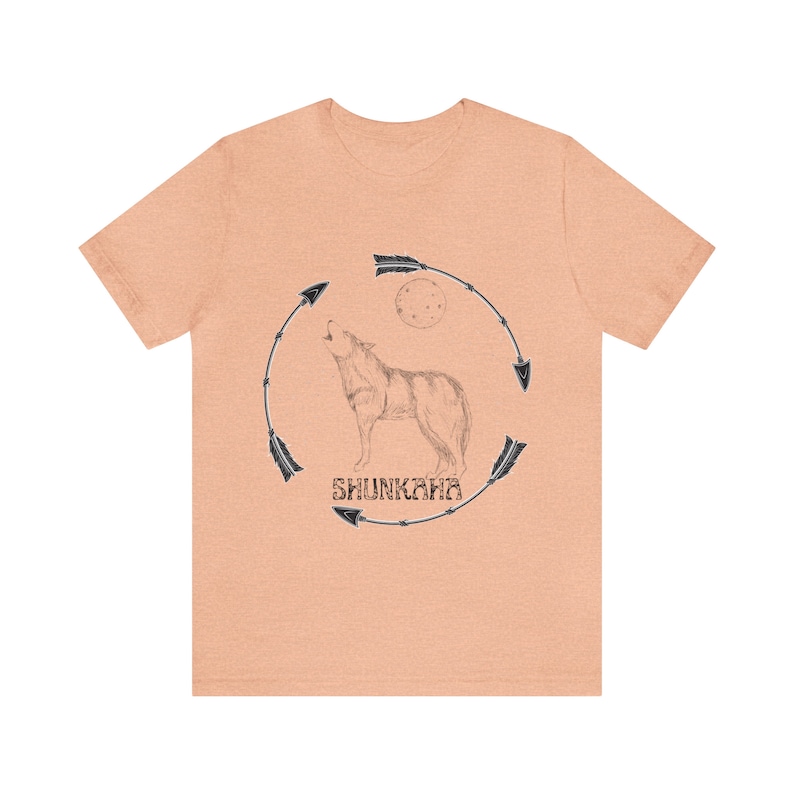 Puede incluir: Una camiseta de color melocot&oacute;n con una ilustraci&oacute;n en blanco y negro de un lobo aullando a la luna. El lobo est&aacute; rodeado por un c&iacute;rculo de flechas con la palabra "SHUNKARA" escrita dentro del c&iacute;rculo.