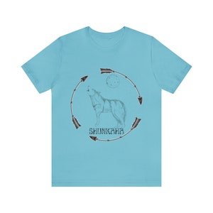 Puede incluir: Una camiseta azul claro con un gr&aacute;fico de un lobo aullando a la luna. El lobo est&aacute; rodeado por un c&iacute;rculo de flechas y la palabra "Shunkara" est&aacute; impresa debajo del lobo.