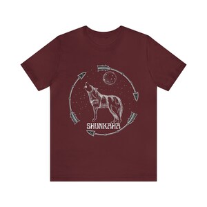 Puede incluir: Una camiseta de color granate con un gr&aacute;fico blanco de un lobo aullando a la luna. El lobo est&aacute; rodeado de flechas y el texto "SHUNKAHA" est&aacute; debajo del lobo.