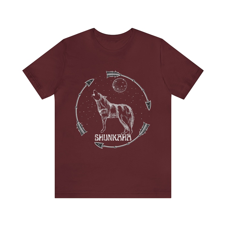 Puede incluir: Una camiseta de color granate con un gr&aacute;fico blanco de un lobo aullando a la luna. El lobo est&aacute; rodeado de flechas y el texto "SHUNKAHA" est&aacute; debajo del lobo.