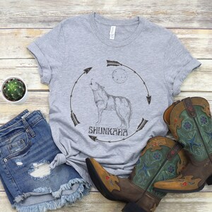 Puede incluir: Camiseta gris con un lobo aullando y el texto "Shunkara" en un c&iacute;rculo con flechas. El lobo est&aacute; aullando a la luna. La camiseta se combina con unos pantalones cortos de mezclilla azul y botas de vaquero marrones y verdes.