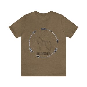 Puede incluir: Una camiseta marr&oacute;n con un lobo aullando a la luna. El lobo est&aacute; rodeado por un c&iacute;rculo de flechas y la palabra "SHUNKARA" est&aacute; escrita debajo del lobo.