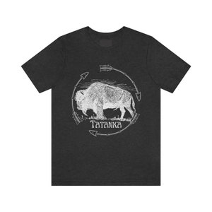 Pu&ograve; includere: Una maglietta grigio scuro con una grafica bianca di un bufalo in un cerchio di frecce. Il testo "Tatanka" &egrave; sotto il bufalo.