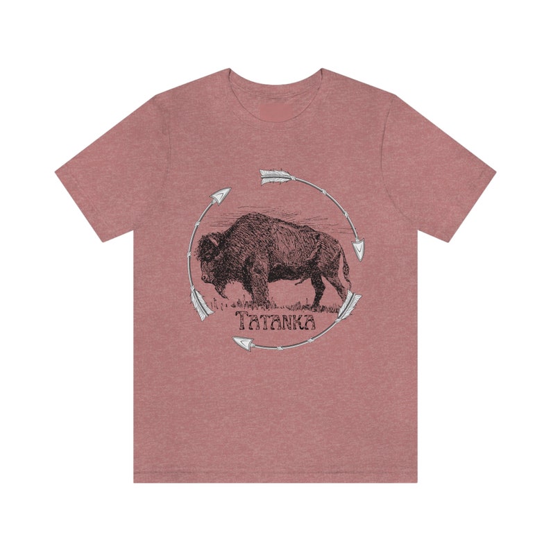 Pu&ograve; includere: Una t-shirt color rosso heather con un'illustrazione in bianco e nero di un bufalo circondato da frecce. Il testo "Tatanka" &egrave; stampato sotto il bufalo.