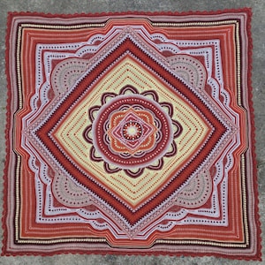 May include: A red, white and cream crochet blanket with a circular design in the centre. The blanket has a scalloped edge and is made with a variety of stitches.