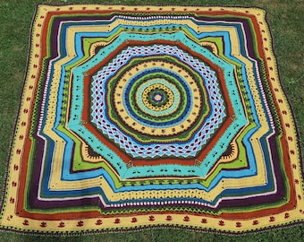 Op de afbeelding: Een kleurrijk gehaakt mandala-deken met een cirkelvormig ontwerp met meerdere lagen van verschillende gekleurde garens. Het deken ligt op een groene grasmat.