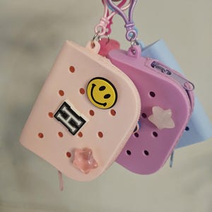 May include: Three small, colorful zippered pouches with a key ring attachment. The pouches are in shades of pink, purple, and blue. The pink pouch has a smiley face and a letter "H" patch. The purple pouch has a star charm. The pouches have a perforated design.