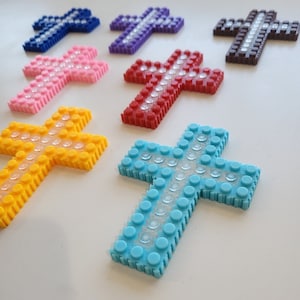 May include: A collection of colorful, pixelated crosses in various hues, including blue, purple, pink, red, yellow, and brown. Each cross is adorned with clear, circular embellishments, creating a sparkling effect. The crosses are arranged on a white surface.