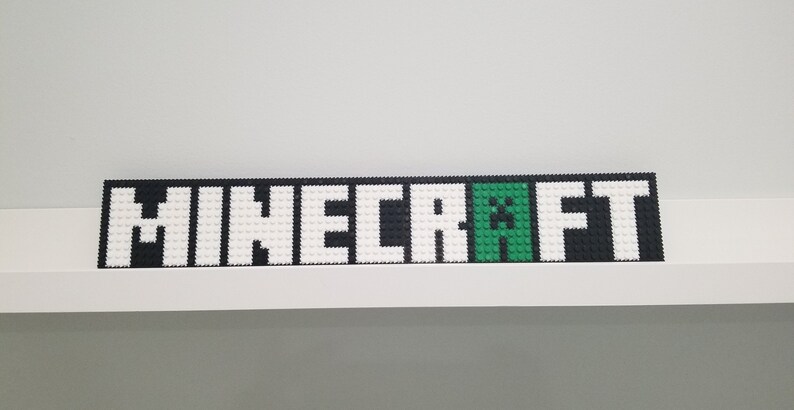 Puede incluir: Un logotipo de Minecraft en blanco y negro con un personaje Creeper verde en el medio. El logotipo est&aacute; hecho de peque&ntilde;os bloques cuadrados.