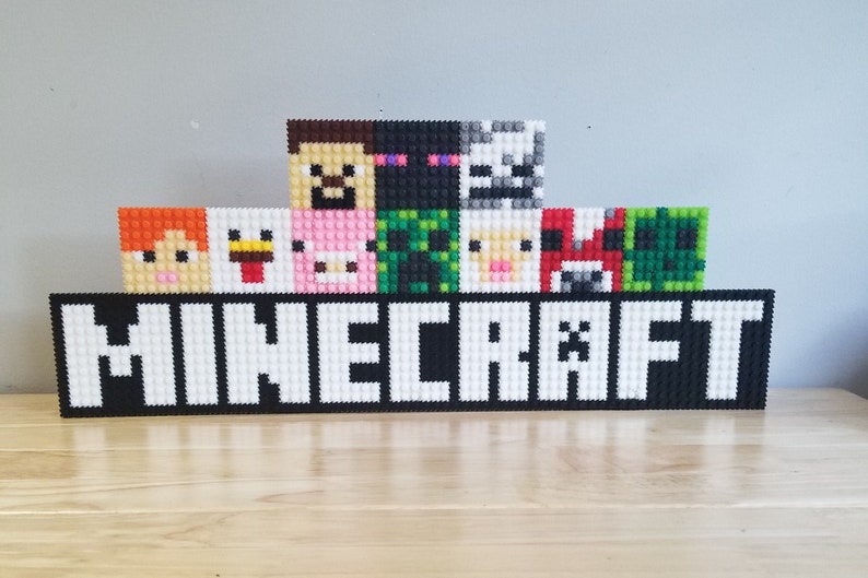 Puede incluir: Una decoraci&oacute;n de pared con tem&aacute;tica de Minecraft hecha de bloques de pl&aacute;stico de colores. La decoraci&oacute;n presenta la palabra "MINECRAFT" en letras blancas sobre un fondo negro, con varios personajes del juego encima.