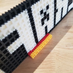Puede incluir: Primer plano de una pieza de arte de p&iacute;xeles en blanco y negro con la palabra "PLAY" escrita. La pieza est&aacute; montada en un marco negro y tiene un soporte rojo y amarillo.