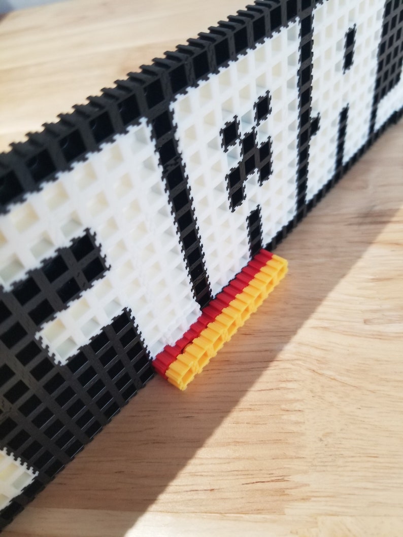 Puede incluir: Primer plano de una pieza de arte de p&iacute;xeles en blanco y negro con la palabra "PLAY" escrita. La pieza est&aacute; montada en un marco negro y tiene un soporte rojo y amarillo.
