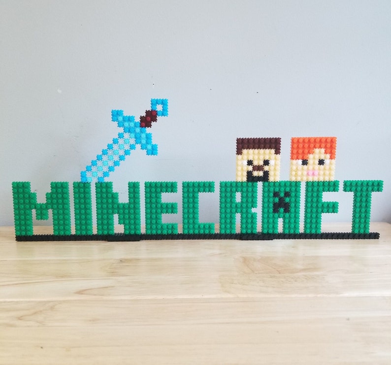 Puede incluir: Un logotipo de Minecraft verde hecho de peque&ntilde;os bloques de pl&aacute;stico con una espada azul y dos personajes pixelados en la parte superior.