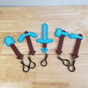 May include: Four keychains featuring Minecraft-inspired pixel art designs. The keychains are made of plastic beads and feature a sword, pickaxe, and two axes. The keychains are attached to black metal key rings.
