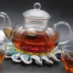 Puede incluir: Una tetera de vidrio con un infusor de vidrio transparente se encuentra sobre un posavasos de ganchillo. La tetera está llena de té marrón y dos tazas de té de vidrio transparente están llenas de té.