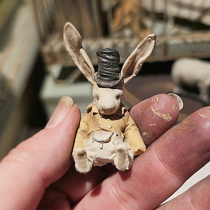 Könnte beinhalten: Eine kleine anthropomorphe Kaninchenfigur mit einem dunkelgrauen Zylinder, einer beigen Jacke mit sichtbaren Knöpfen und einer hellen Hose. Das Kaninchen hat lange, aufrechte Ohren und eine silberfarbene Kette an der Taille der Hose.