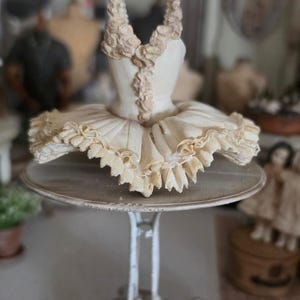 Könnte beinhalten: Ein cremefarbenes Balletttutu mit strukturiertem Oberteil und gerüschtem Rock, präsentiert auf einem weißen Metallständer. Das Oberteil zeigt komplizierte, erhabene blumenartige Details entlang des Ausschnitts und in der Mitte, mit einem Rock aus geschichtetem, plissiertem Stoff.