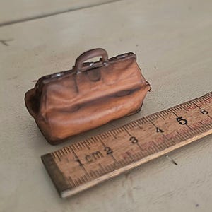 Könnte beinhalten: Eine braune Miniatur-Arzttasche mit einem einzelnen Tragegriff und einem strukturierten Rahmen. Die Tasche liegt neben einem Holzlineal, das zeigt, dass der Gegenstand eine Länge von etwa 6,5 cm von Ende zu Ende aufweist.