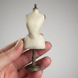 Peut inclure: Un petit mannequin de couturière crème sur un support vert foncé. Le torse est détaillé avec une forme incurvée et un petit fleuron sombre en haut. La base est ronde et le support est orné.