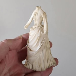 May include: A cream-colored miniature figurine of a dress with long sleeves, a high collar, and a fitted bodice. The skirt features intricate draped fabric detailing and vertical pleats at the hem, capturing a structured, historical garment design.