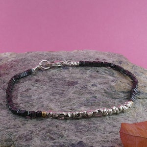 May include: A black hematite beaded bracelet with silver and gold accents. The bracelet has a silver clasp and is shown on a gray rock.