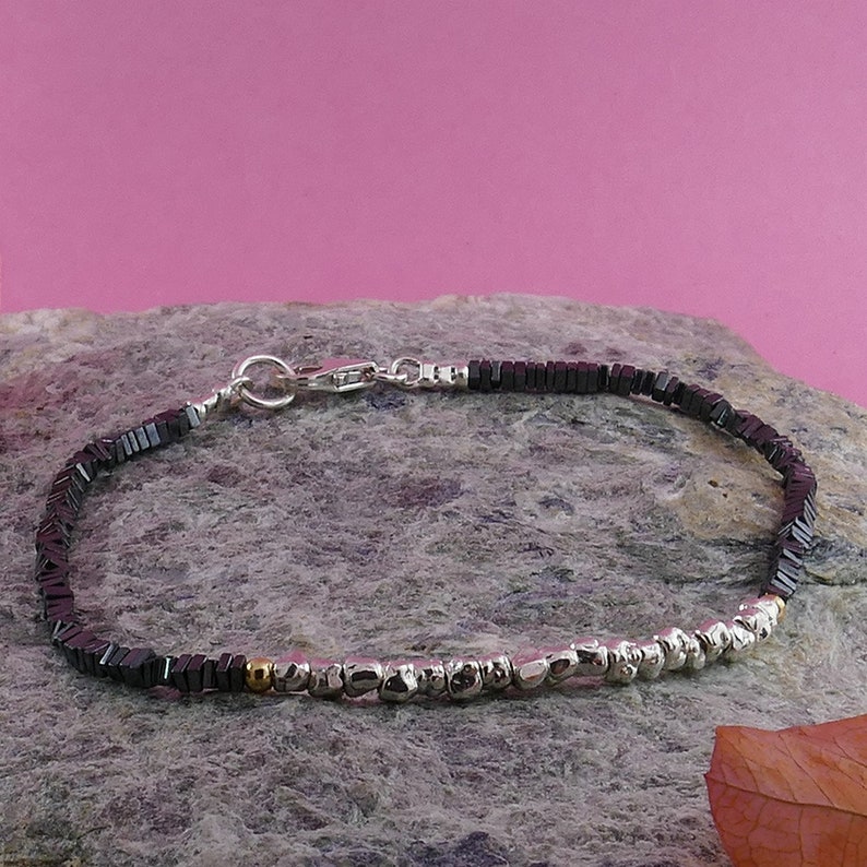 May include: A black hematite beaded bracelet with silver and gold accents. The bracelet has a silver clasp and is shown on a gray rock.