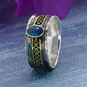 Peut inclure: Une bague en argent et or avec une pierre précieuse en opale bleue. La bague a une bande texturée avec une incrustation en or.