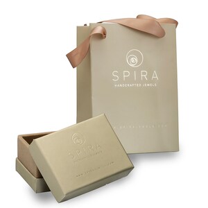 May include: A beige gift bag with a white logo that says "SPIRA HANDCRAFTED JEWELS" and a website address. The bag has a matching beige ribbon handle. A smaller beige box with the same logo is open and sitting in front of the bag.