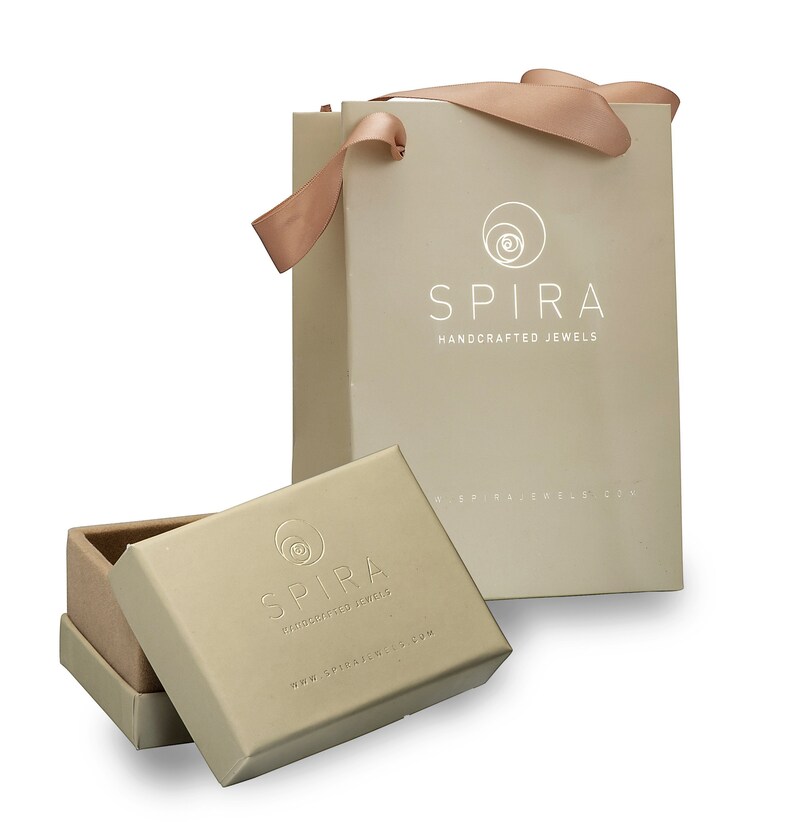 May include: A beige gift bag with a white logo that says "SPIRA HANDCRAFTED JEWELS" and a website address. The bag has a matching beige ribbon handle. A smaller beige box with the same logo is open and sitting in front of the bag.