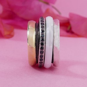 May include: A set of four stacking rings. The rings are made of different materials, including gold, silver, and black onyx. The rings are stacked on top of each other, with the gold ring on the bottom, followed by the silver ring, the black onyx ring, and the white ring on top.