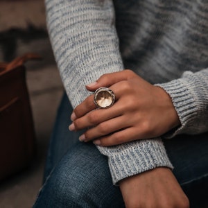 Peut inclure: Une bague en argent avec un grand centre rond en or martelé. La bague est portée à l'index d'une main.
