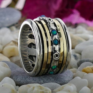 Può includere: Un anello rotante in argento e oro con una fascia di opali verdi. L'anello ha una superficie testurizzata ed è posto su un letto di pietre bianche.