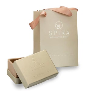 May include: A beige gift bag with a pink ribbon and the text "SPIRA HANDCRAFTED JEWELS" printed on the front. The bag is sitting on a white surface next to a beige box with the same logo and text.