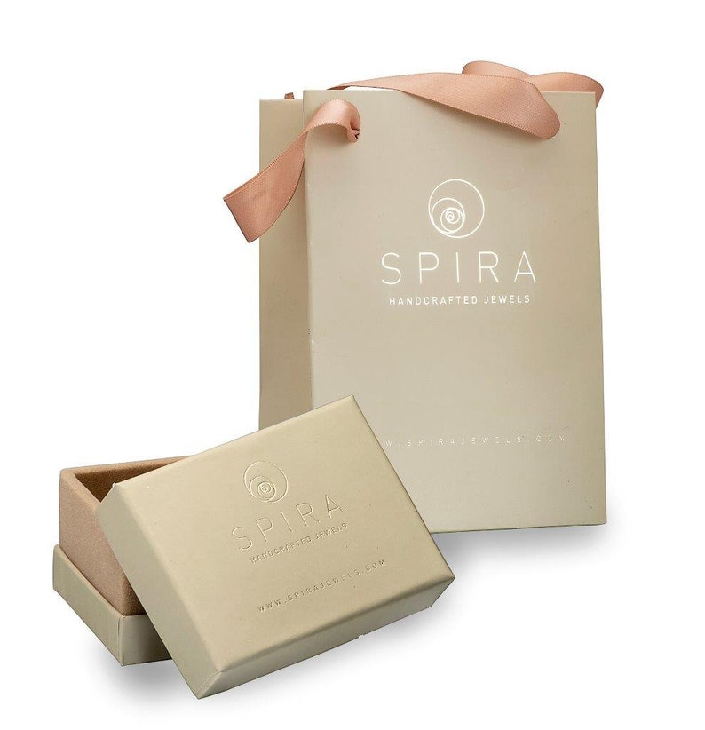 May include: A beige gift bag with a pink ribbon and the text "SPIRA HANDCRAFTED JEWELS" printed on the front. The bag is sitting on a white surface next to a beige box with the same logo and text.