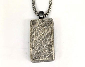May include: A rectangular pendant with a textured, scratched surface hangs from a silver chain. The pendant is a muted silver color, and the chain has a small, round link design. The necklace is displayed against a white background.
