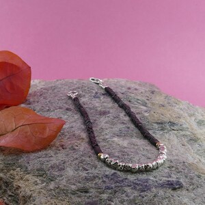 May include: A black beaded bracelet with silver and gold accents. The bracelet is on a gray rock with a pink background.
