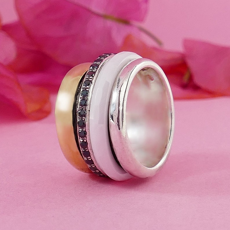 May include: A multi-layered ring with a white enamel band, a gold band, and a band of black gemstones. The ring is made of silver and has a polished finish.