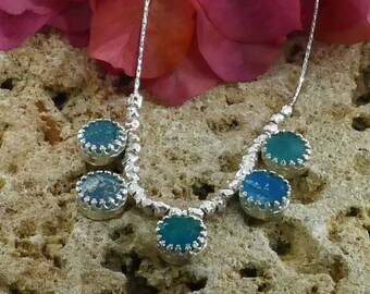 May include: A silver necklace with five round blue and green glass stones set in silver bezels. The necklace is on a rocky surface with pink flower petals in the background.
