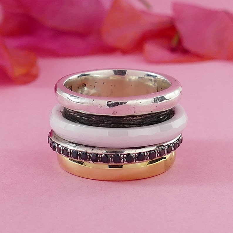 May include: A stack of four rings, one silver, one white, one black with black gemstones, and one gold. The rings are stacked on top of each other.