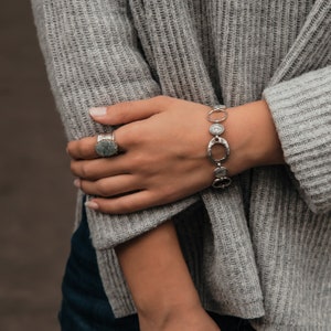 May include: A silver bracelet with oval and round links, featuring a textured finish. The bracelet is worn on a person's wrist, along with a silver ring with a green stone.