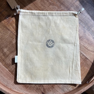 Peut inclure: Un sac beige à cordon de serrage avec un logo circulaire indiquant "East Wind Trading Post" autour d'une étoile. Le sac est fait d'un tissu texturé et possède une petite étiquette attachée. Le sac est posé sur une surface en bois.