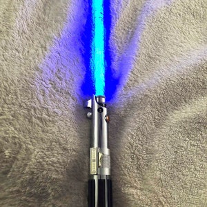May include: A blue lightsaber with a black handle and silver accents. The lightsaber is turned on and emitting a blue light.