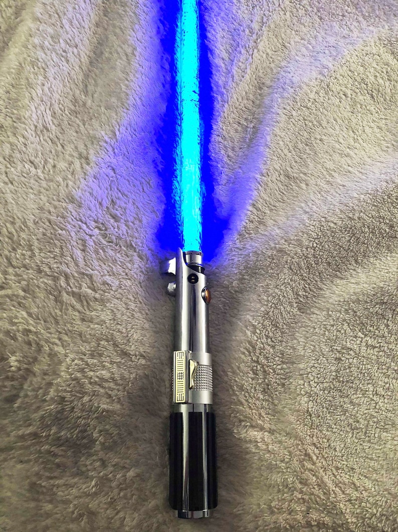 May include: A blue lightsaber with a black handle and silver accents. The lightsaber is turned on and emitting a blue light.