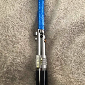 May include: A silver and black lightsaber toy with a blue light-up blade. The lightsaber has a gold accent and a textured grip.