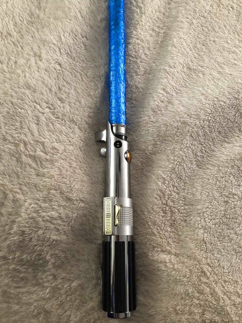 May include: A silver and black lightsaber toy with a blue light-up blade. The lightsaber has a gold accent and a textured grip.