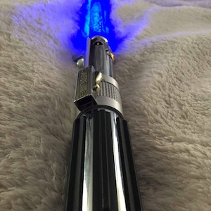 May include: A silver and black lightsaber with a blue blade. The lightsaber is on a white fuzzy surface.