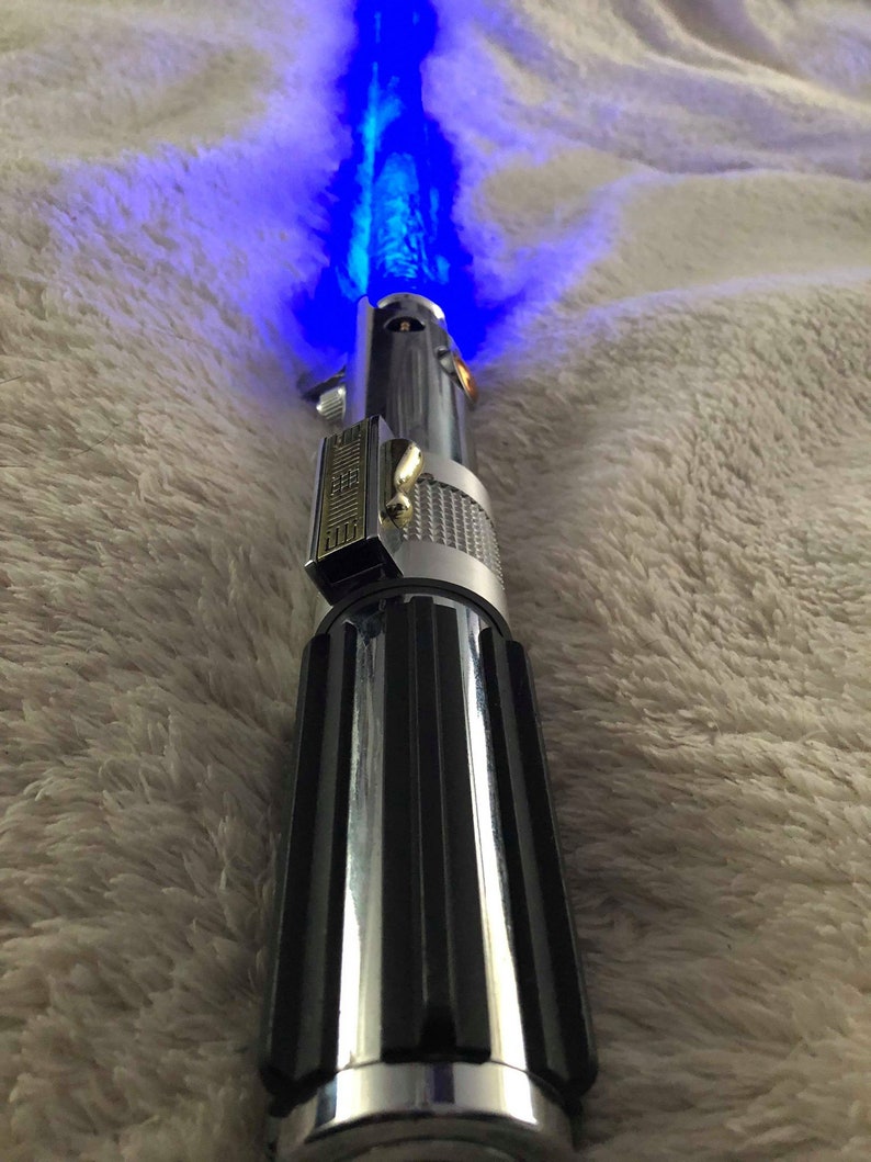 May include: A silver and black lightsaber with a blue blade. The lightsaber is on a white fuzzy surface.