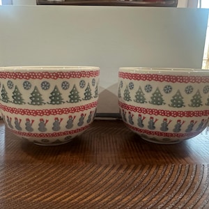 May include: Two ceramic mugs with a festive winter design. The mugs feature a white base with a red band of dots and a band of snowmen. Above the red band are green Christmas trees and snowflakes. Each mug has a white handle.