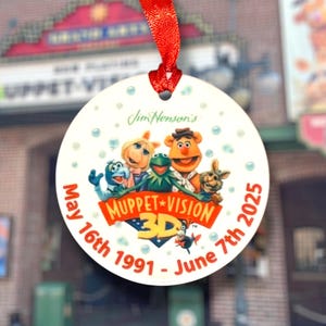 May include: A round, white ornament with a red ribbon. The ornament features the Muppet characters, the text "Muppet Vision 3D," and the dates "May 16th 1991 - June 7th 2025." The ornament is decorated with blue bubbles.